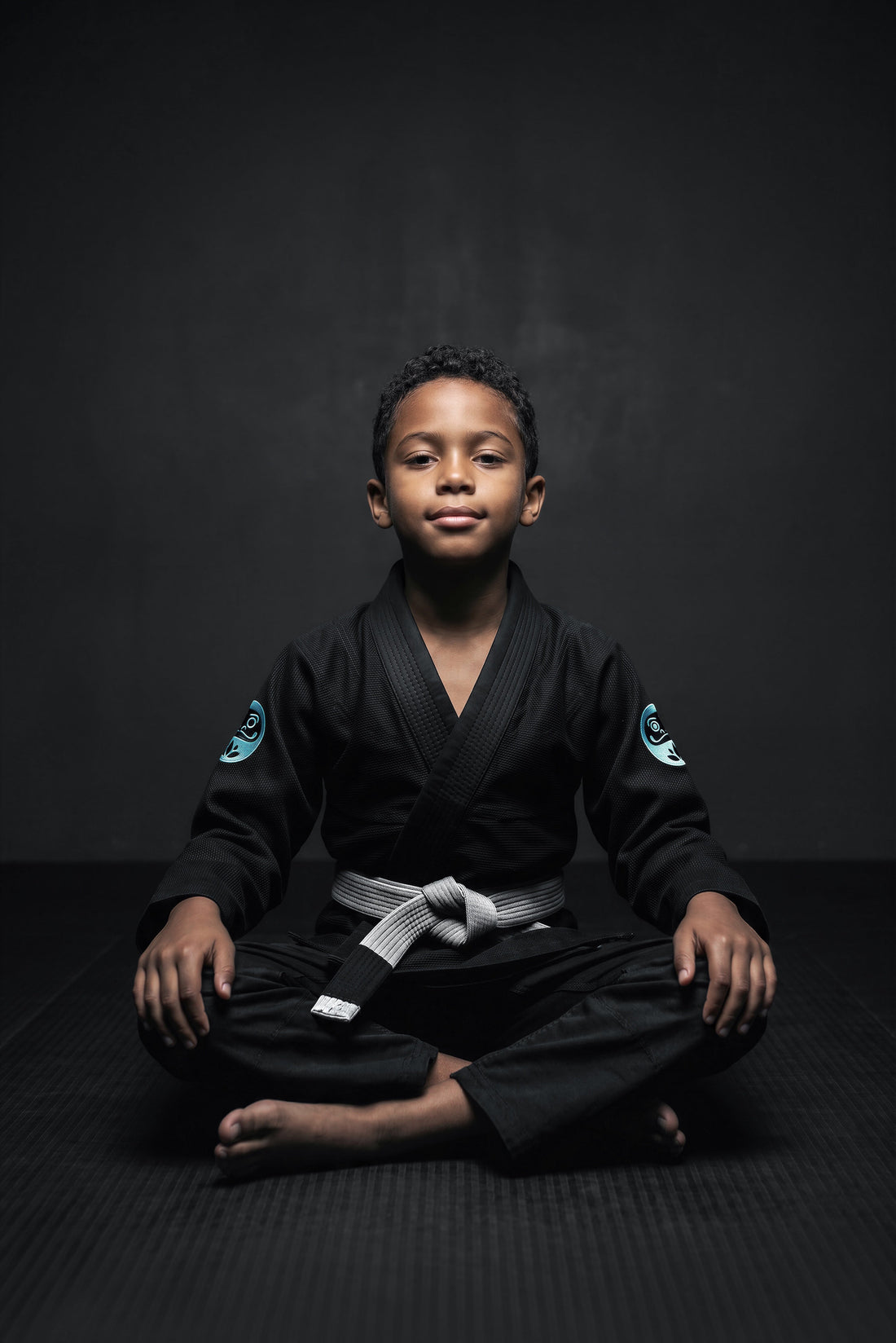 Child in a black Daruma Kodomo organic cotton BJJ Gi, seated on the mats