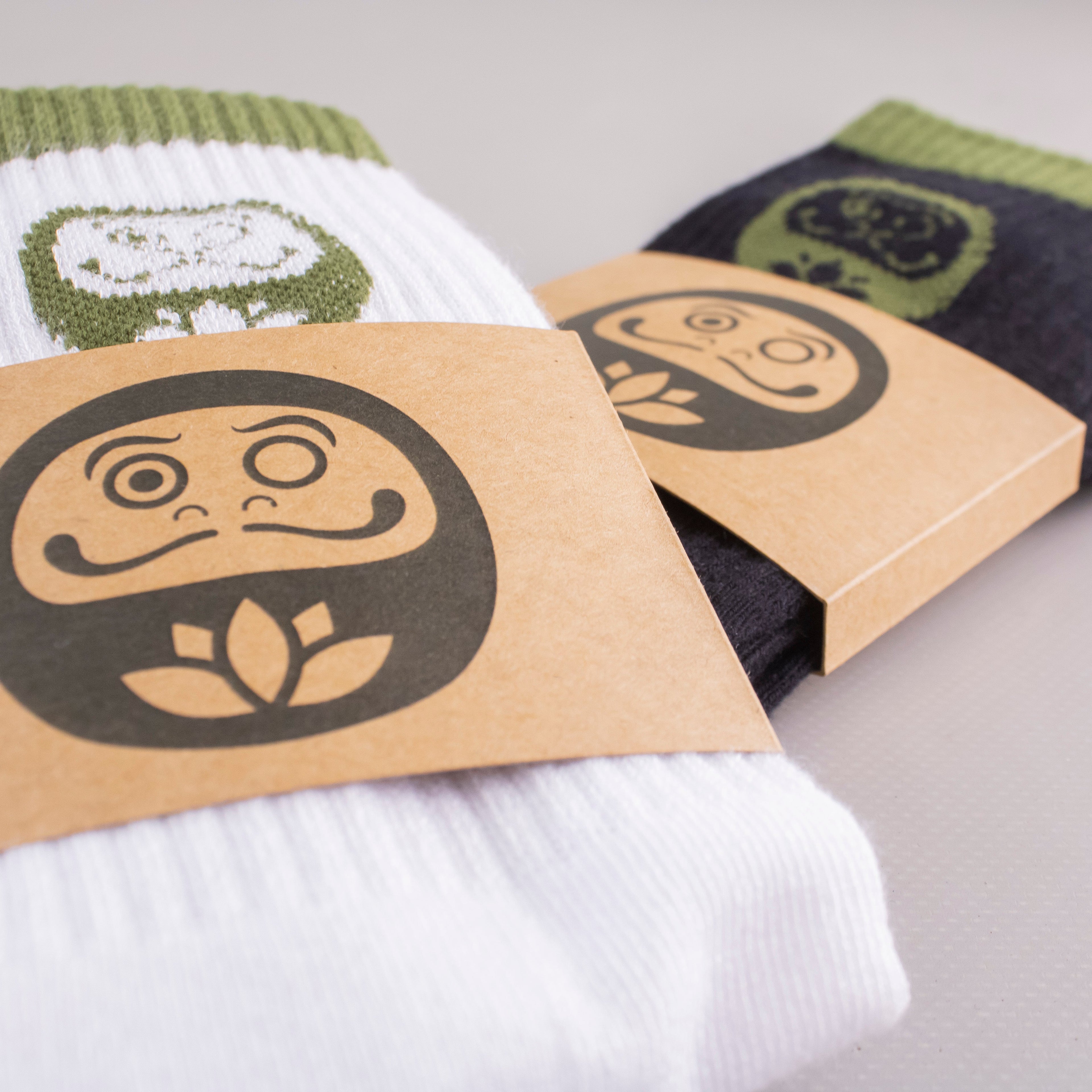 Pair of socks with a Daruma design on a cardboard packaging label, on a light gray background.